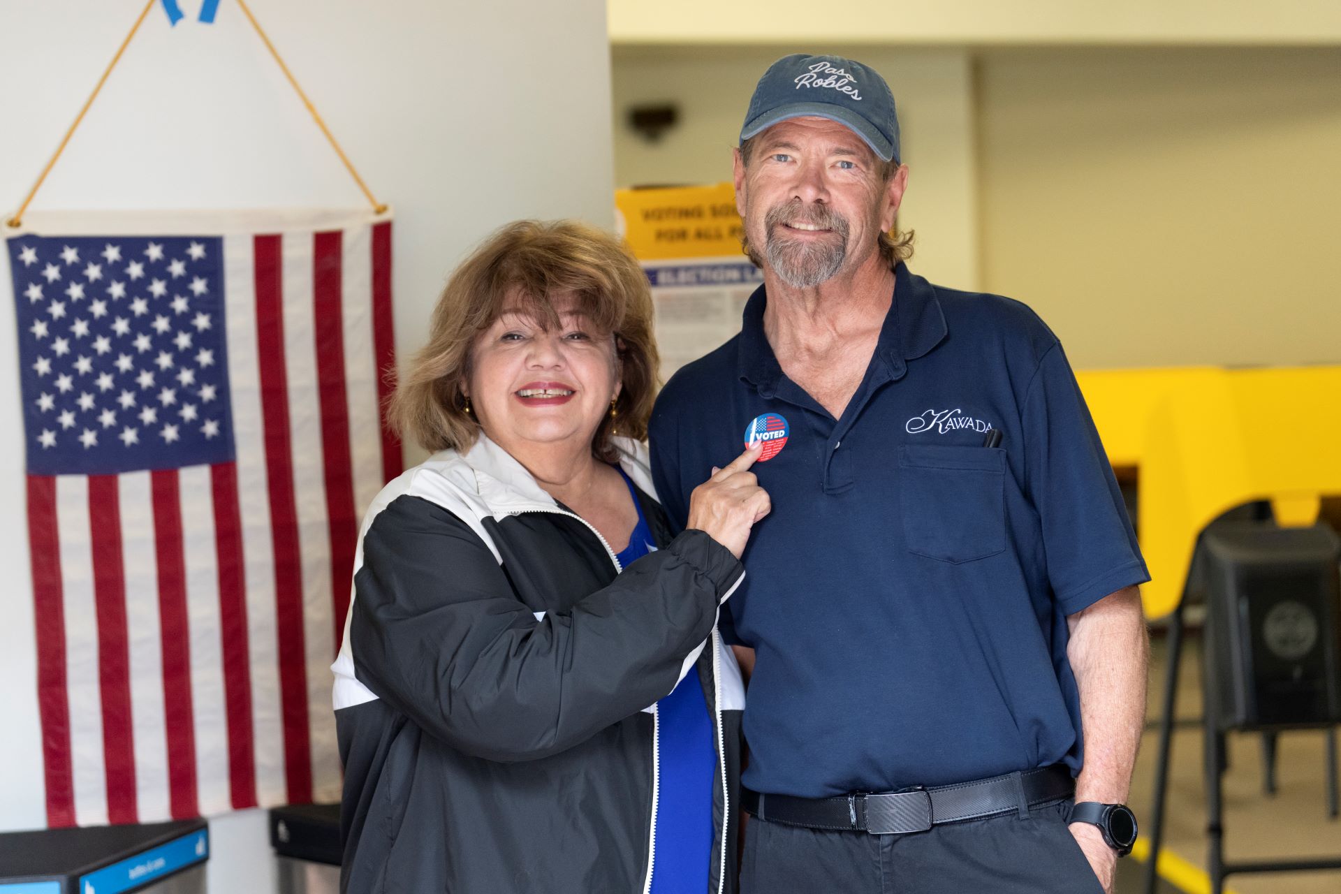 Female and male standing together with the woman holding an "I Voted" sticker, United States flag in the background and voting booths