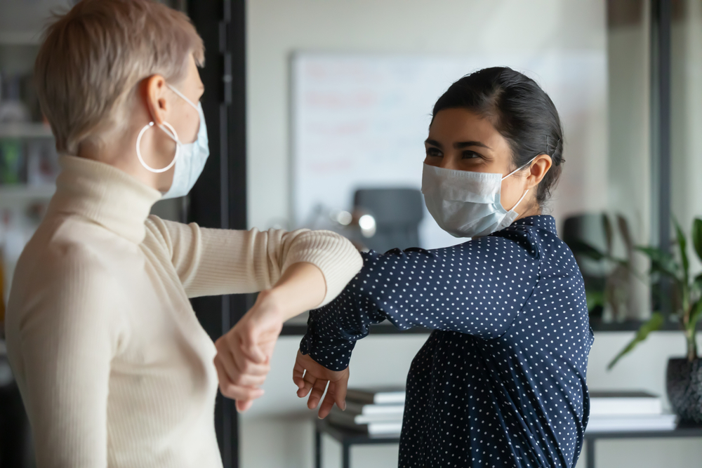 A smiling woman elbow bumps her colleague while wearing a face mask.