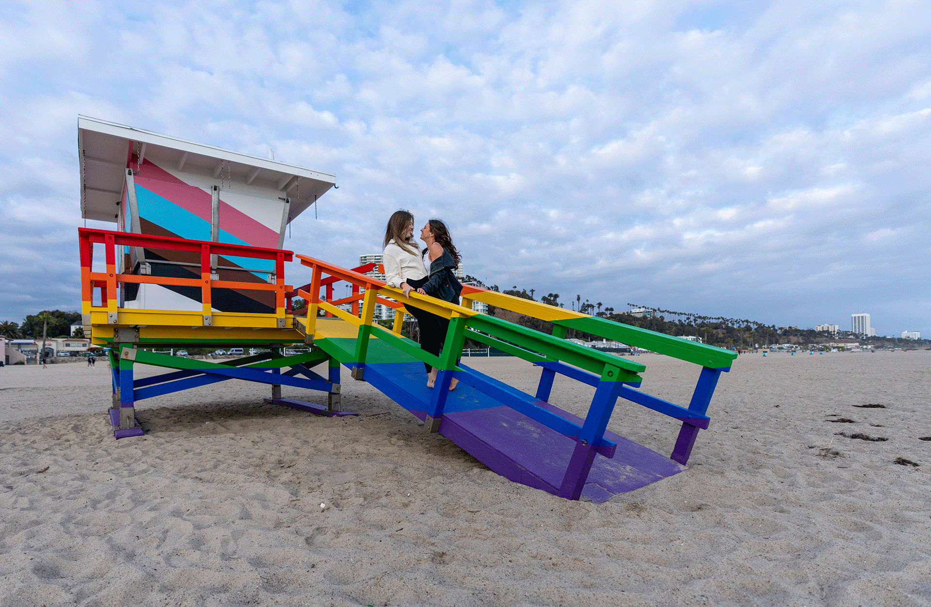 An LGBTQ+ couple embraces on a lifeguard tower painted in Pride colors at Ginger Rogers beach.