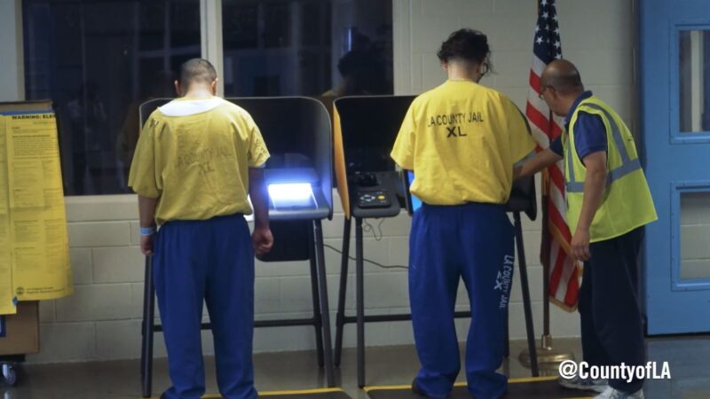 Two men wearing a yellow shirt and blue pants looking towards voting machine with worker next to one of them as the cast their votes at Twin Towers correctional facility