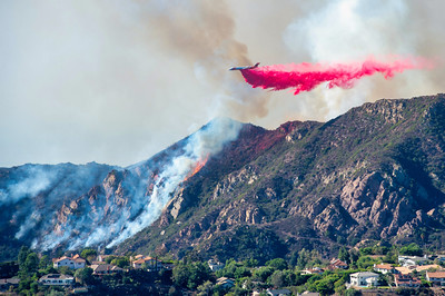 Firefighting aircraft drops red fire retardant over a brush fire on a hillside near homes, with smoke rising in the background.