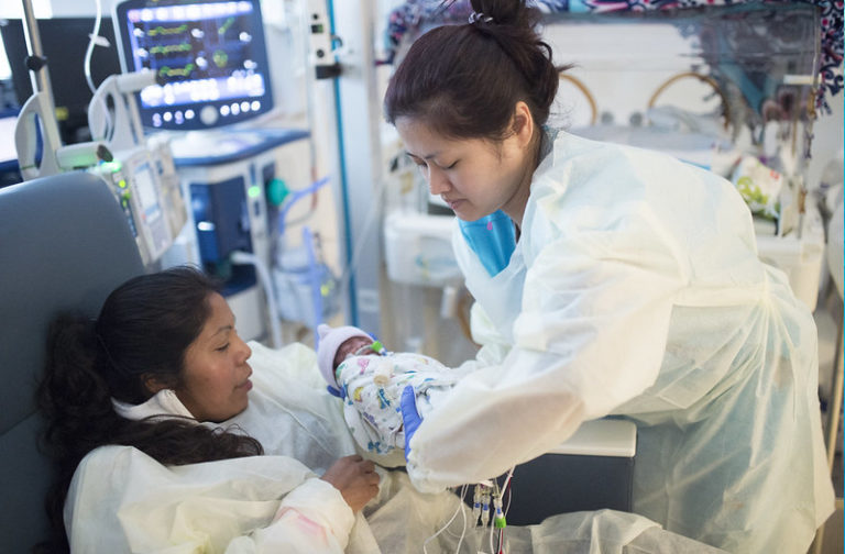 A healthcare worker in protective clothing cares for a newborn baby while the mother rests in a hospital bed, with medical monitors visible in the background.