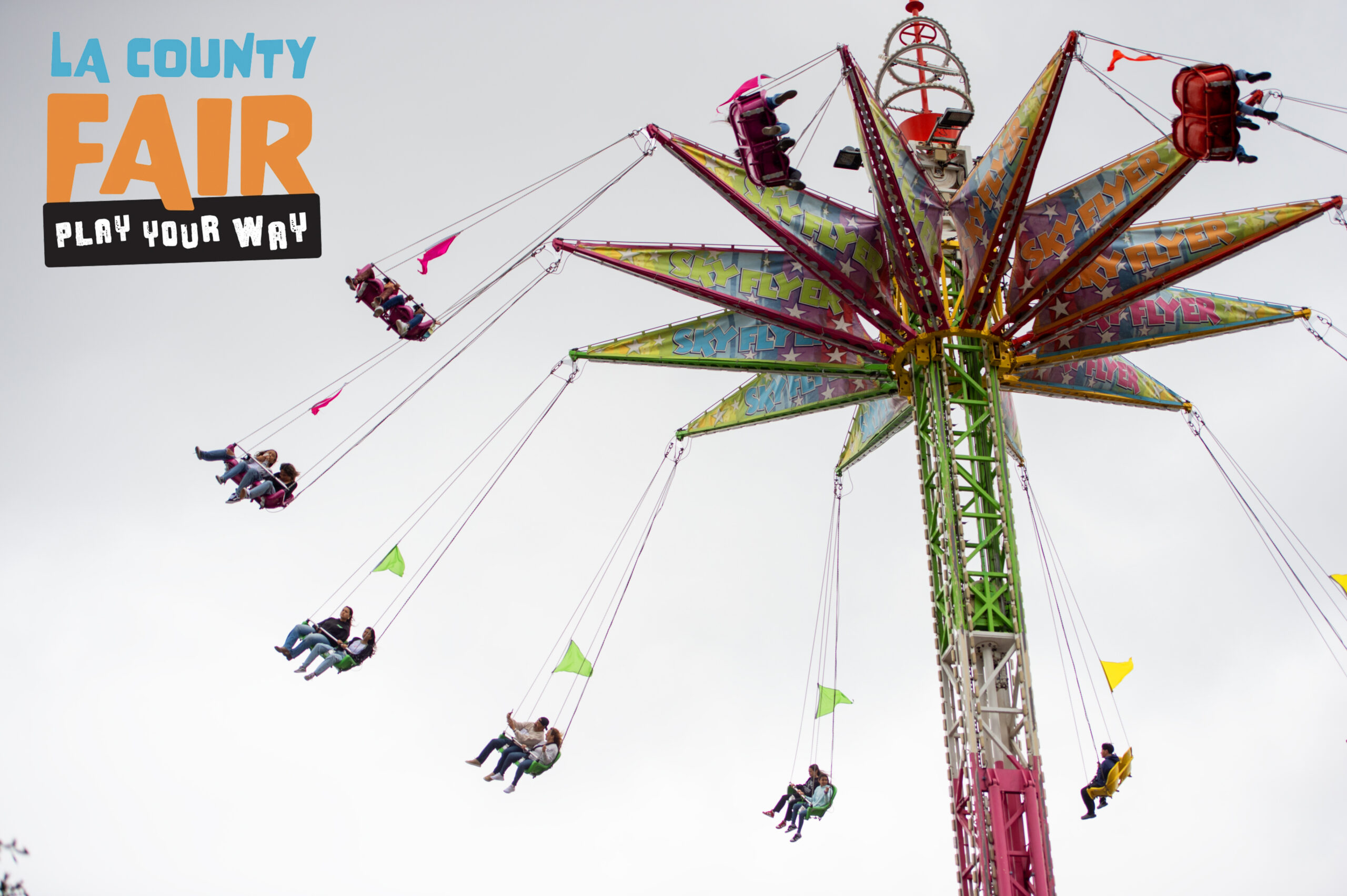 People riding a colorful swing carousel ride at the LA County Fair, suspended high in the air against a light sky, with ‘LA County Fair – Play Your Way’ text displayed on the left.