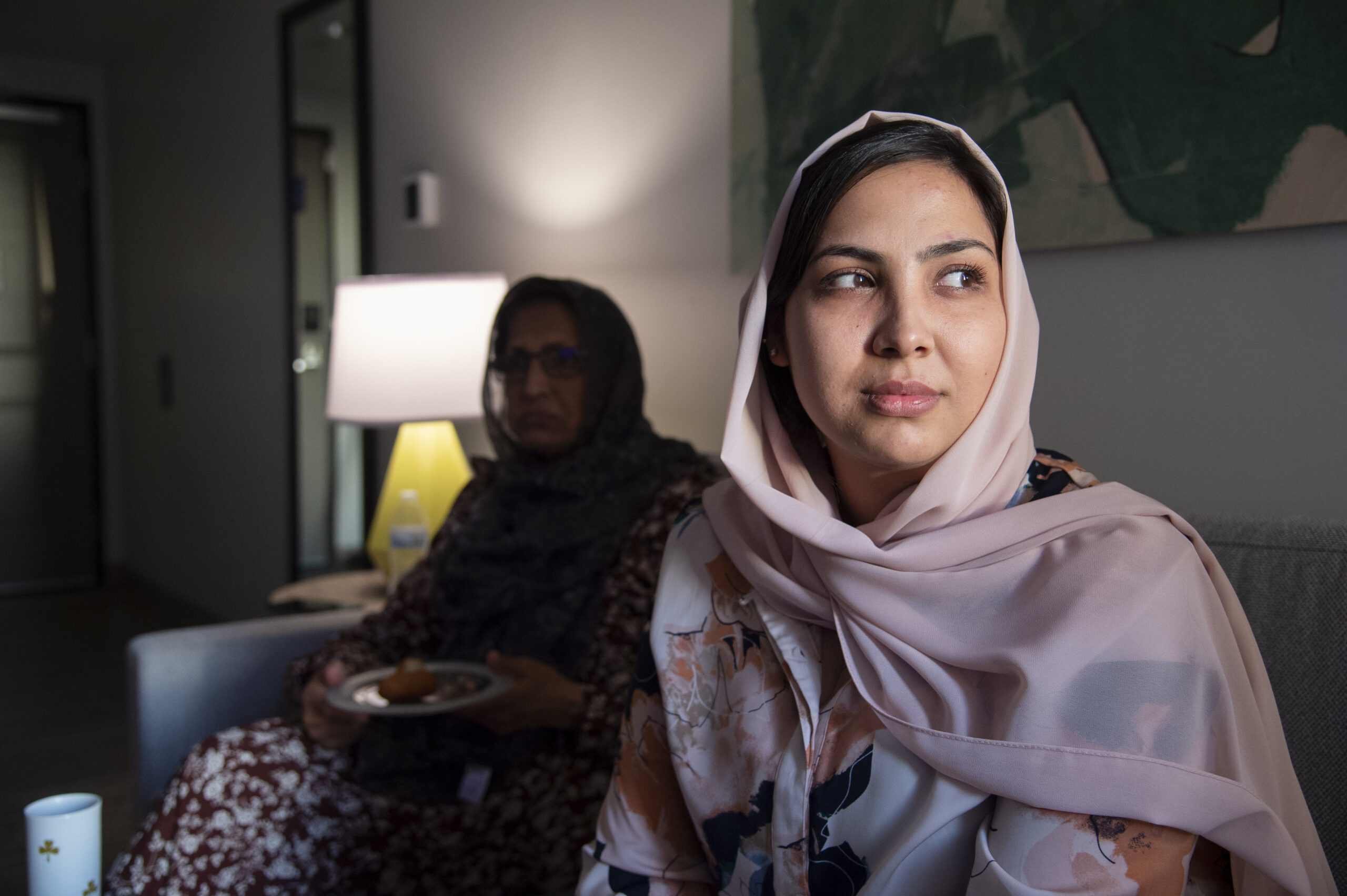 A woman wearing a light pink headscarf sits indoors, looking thoughtfully off to the side. Another woman sits blurred in the background near a softly lit lamp, creating a quiet, reflective atmosphere